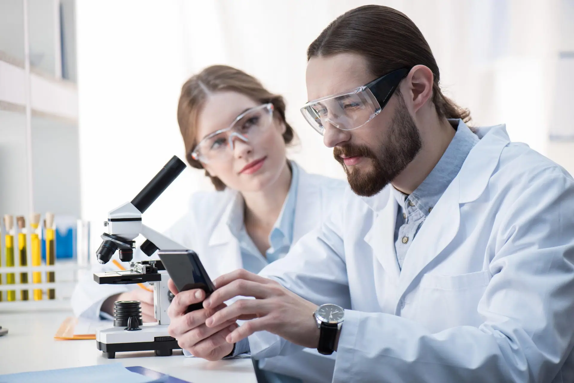 Two scientists in lab coats using a smartphone next to a microscope, demonstrating the PicoTeam Mobile App for accessing Digital Cage Card data.